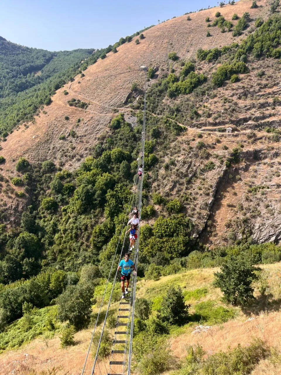 People crossing a long suspended rope bridge over a green valley