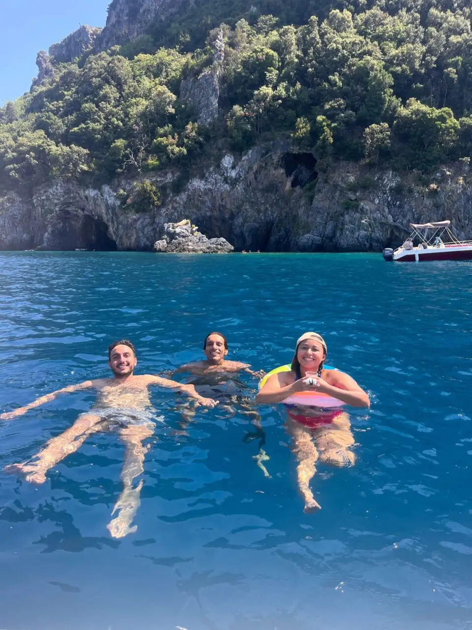 Three friends swimming together in clear turquoise sea near a rocky coastline