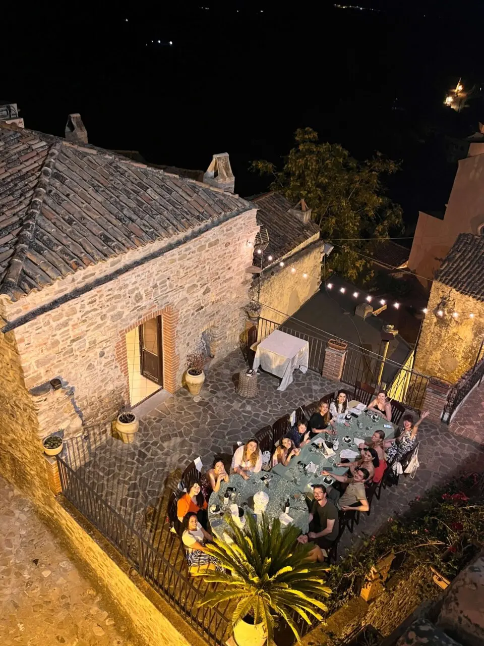 Outdoor dinner gathering on a stone terrace at night with string lights
